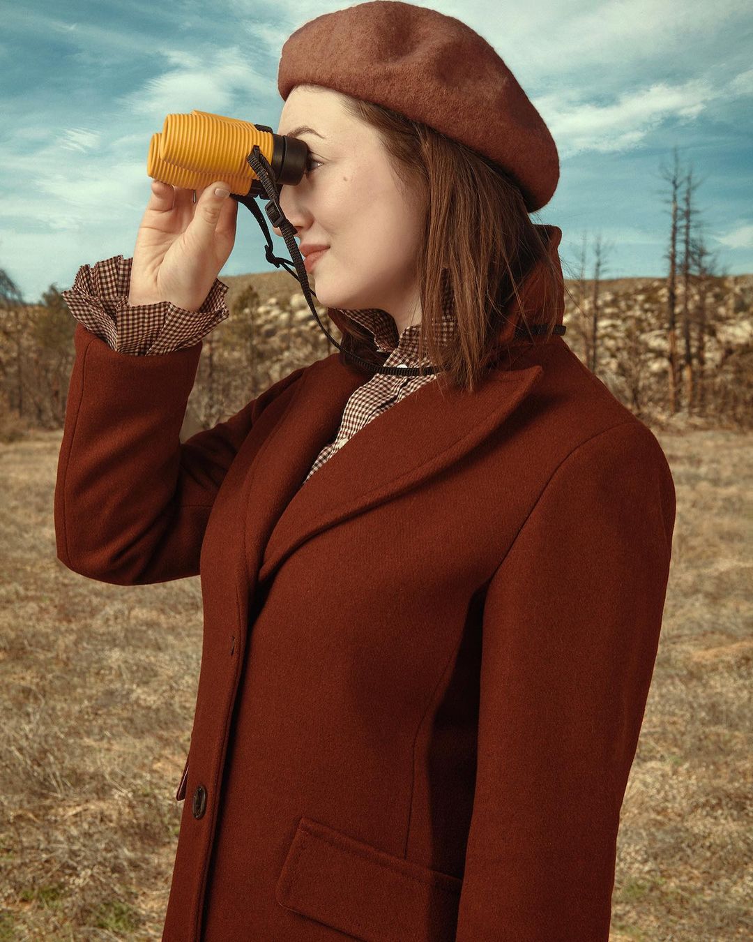 A woman in a beret and coat looks through yellow binoculars in a grassy field with bare trees and a blue sky, vintage-style photography.